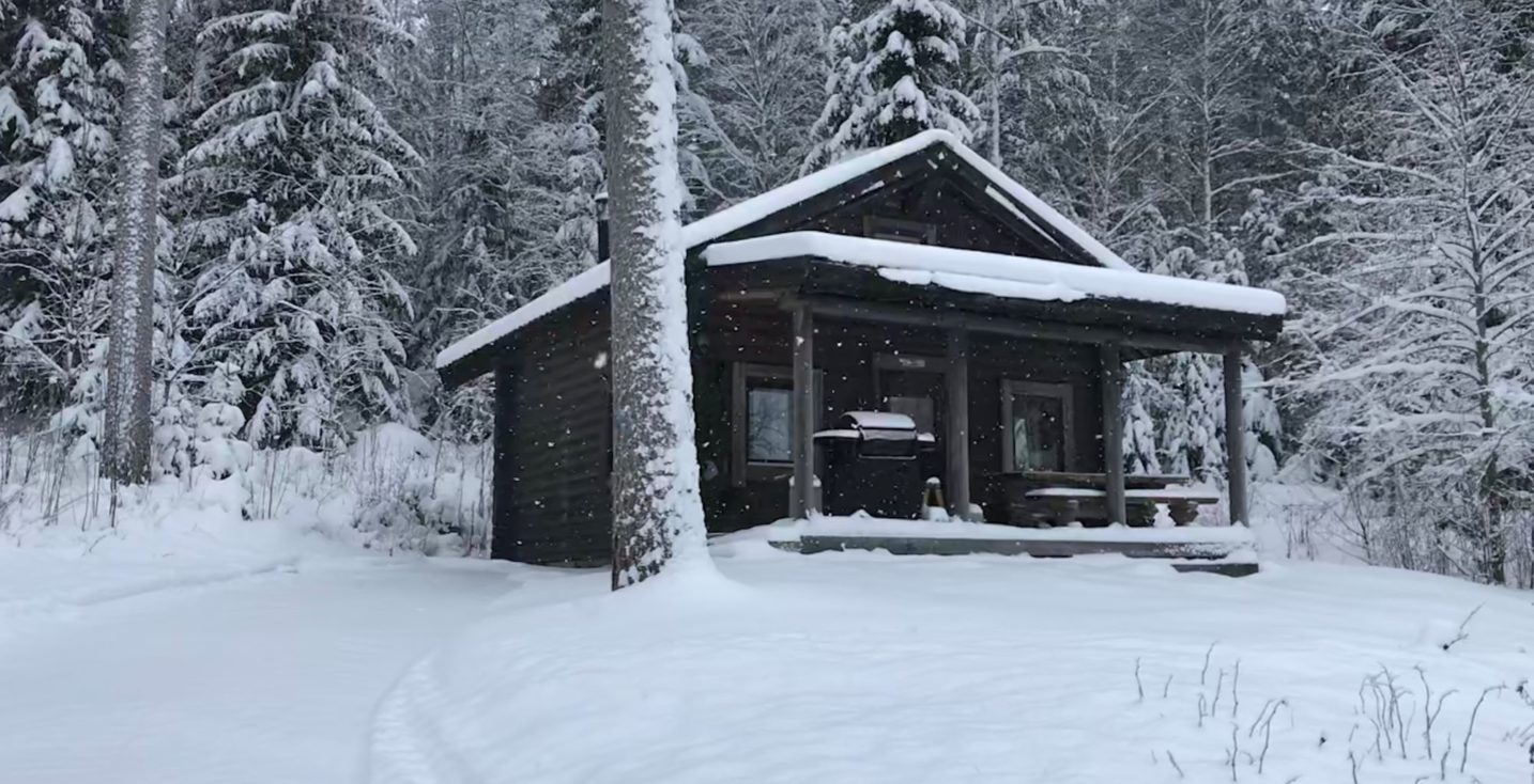 Aino-torppa talvimaisemissa lumihiutaleiden leijuessa.